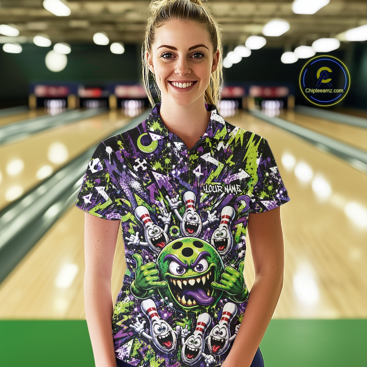 Front view of a female model at a bowling lane wearing a green and purple bowling polo shirt featuring a mischievous green monster bowling ball with sharp teeth, a purple tongue, playful hand gesture, and cheerful cartoon bowling pins.