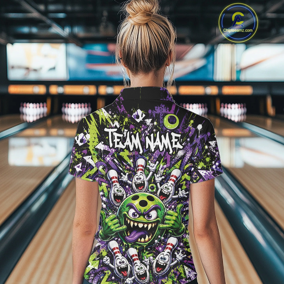 Back view of a female model in a bowling lane wearing a green and purple bowling polo shirt showcasing a mischievous green monster bowling ball with sharp teeth, a purple tongue, playful hand gesture, and cheerful cartoon bowling pins.