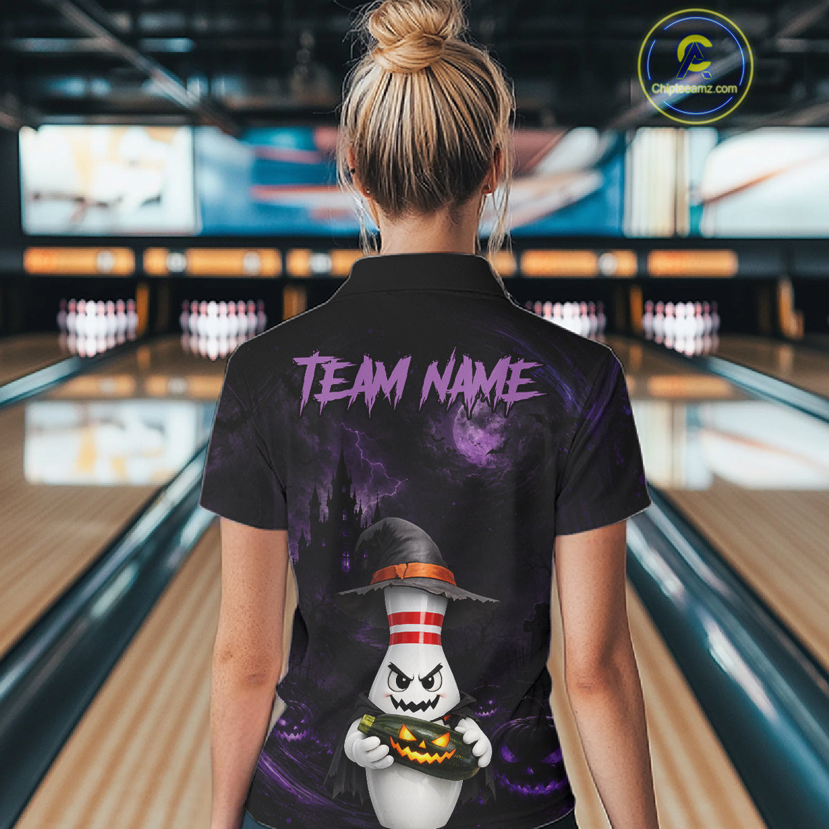 A woman viewed from behind wearing a black short-sleeve polo shirt with a Halloween-themed graphic print. The shirt features purple 'TEAM NAME' text and a cartoon ghost wearing a witch hat holding a pumpkin, set against a bowling alley background.