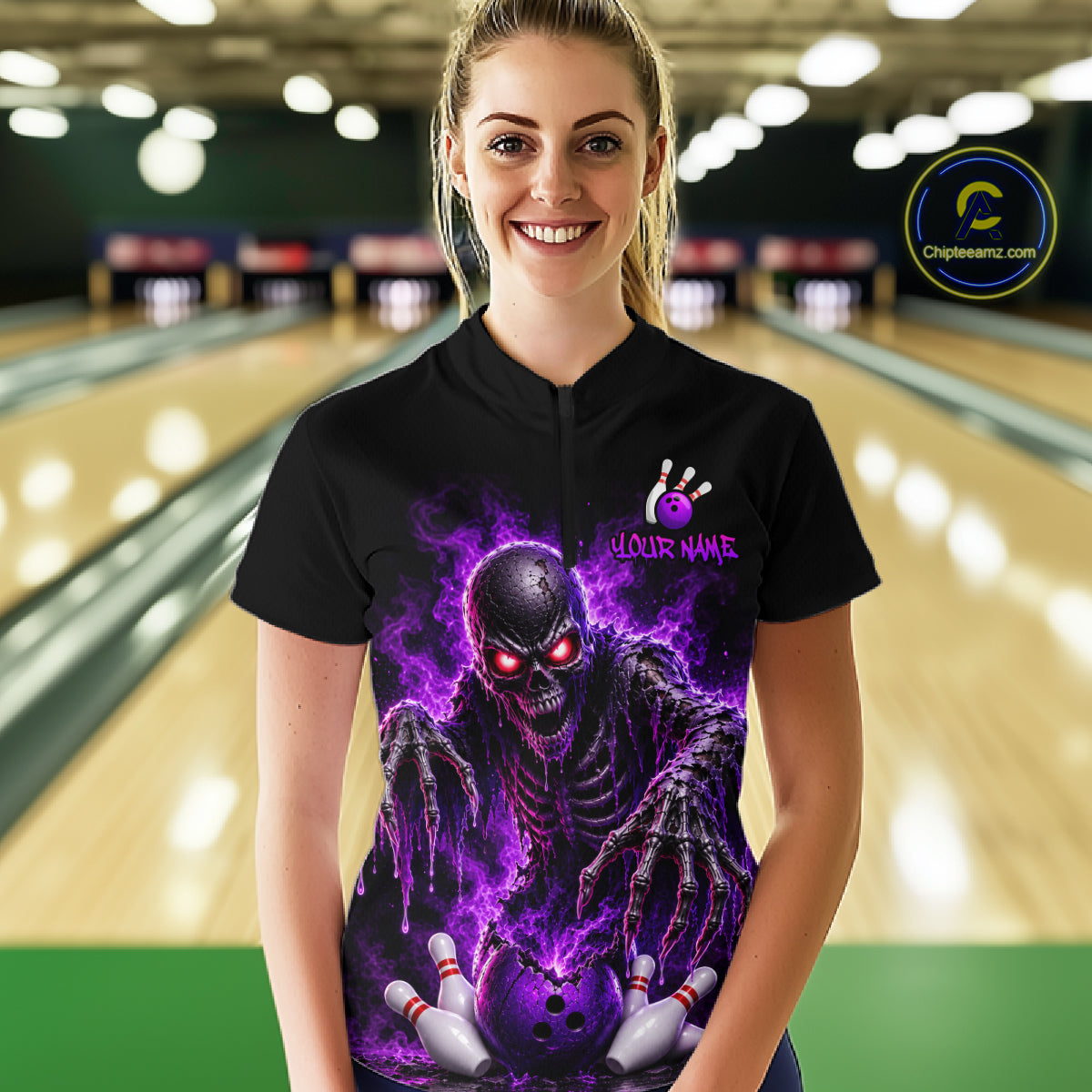 A woman stands in a bowling alley wearing a black short-sleeve bowling shirt with a large purple and black skull graphic on the front, holding bowling pins.